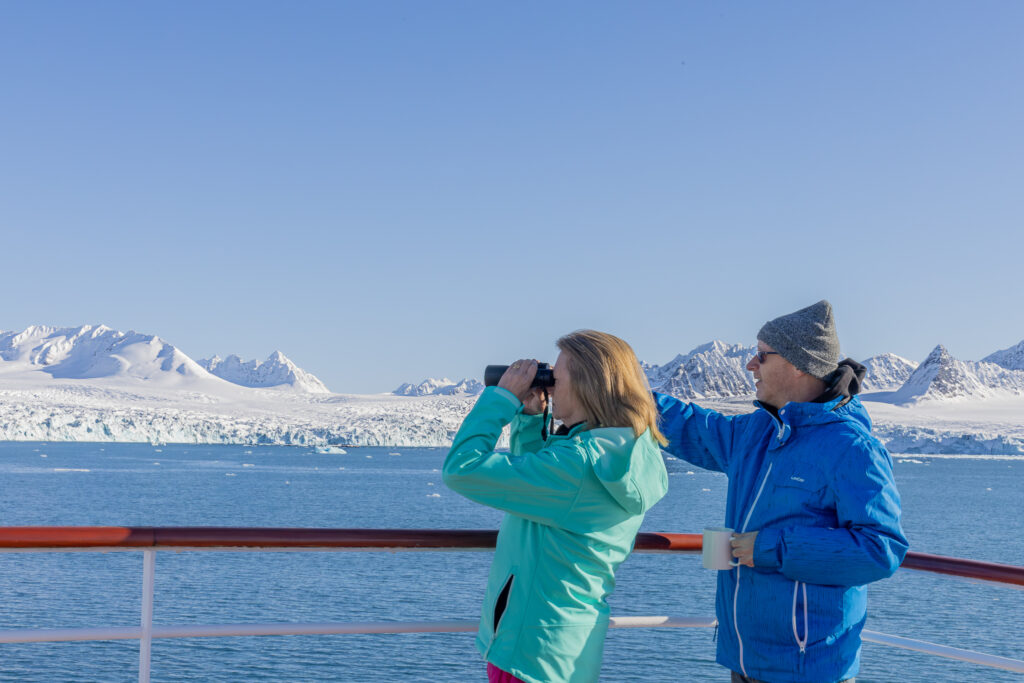 couple looking out to see from a cruise ship, pointing to something in the distance with snow covered mountains in the backgroun
